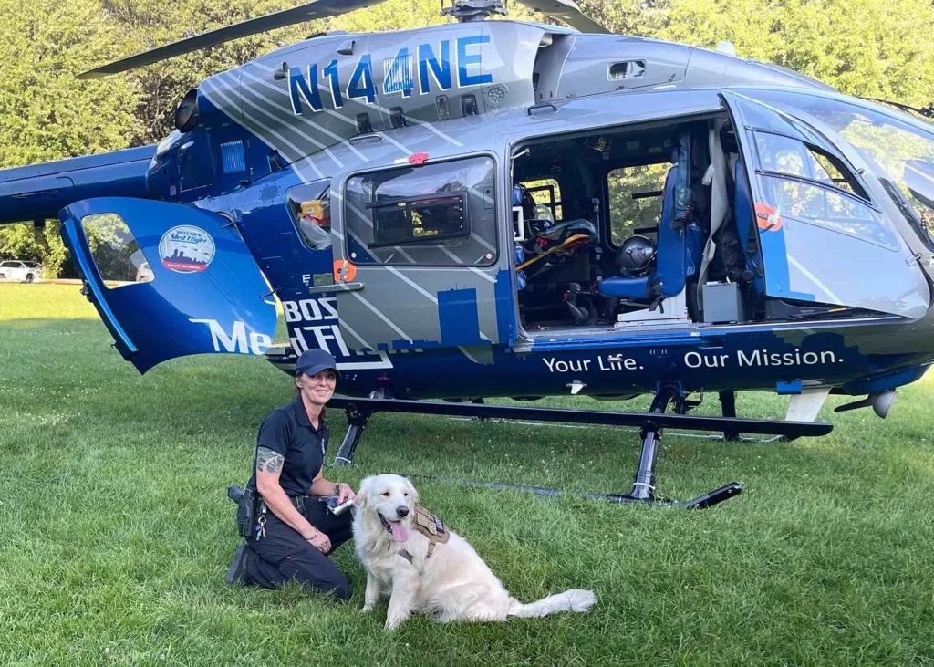 Harper lays in grass in front of a blue and silver rescue helicopter with Detective Kristen Gagnon.